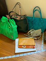 Six purses and bags arranged on floor including large turquoise woven tote, green fabric tote, floral fabric bag, black leather shoulder bag, patterned small crossbody bag, and two wallets.