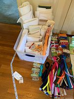 Photo showing white/off-white fabric fat squares and rolls arranged in a plastic bin alongside assorted colorful sewing zippers and small sewing accessories on the floor.