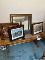 Five framed pieces of wall art on a bench showing various scenes including two fox hunting prints and others.