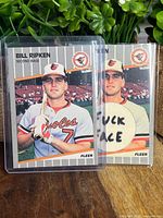 Two Billy Ripken 1989 Fleer baseball cards in protective sleeves; one card shows standard 1989 Fleer design, the other shows a custom art card with explicit text on the bat knob.