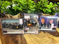 Three Formula One trading cards featuring Max Verstappen and Lewis Hamilton shown on a wooden table with greenery background.