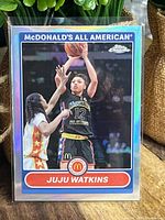 Front of Juju Watkins basketball card from McDonald's All American set showing player in black uniform shooting a basketball with opponent in red and yellow.