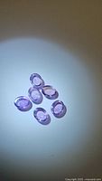 Six oval cabochon natural amethyst gemstones on white background under light showing translucent purple color and polished domed surfaces.