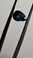 Close-up photo of pear cut natural blue sapphire held by tweezers showing its deep blue color and internal facets under light.
