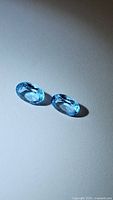 Pair of two oval faceted Swiss blue topaz gemstones placed on a flat surface with light reflecting off their facets.