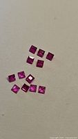 Photo showing 13 small square cut rubies spread on a plain light background, displaying their pink reddish color and transparency.