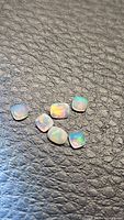 Six natural faceted cushion cut Ethiopian crystal opals on black textured surface. Stones exhibit colorful flashes of orange, green and blue.