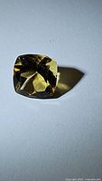 Close-up photo of a cushion cut golden citrine gemstone on a light grey surface showing facets and clarity.