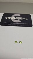 Pair of small oval cut apple green peridot gemstones on white surface with GRWN Gems business card.
