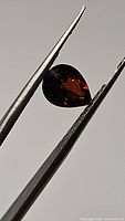 Close-up photo of a pear-shaped deep orange-red tourmaline gemstone held by tweezers, showing faceted surfaces and internal reflections.