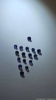 Top-down view of 17 natural blue sapphire diamond cut gemstones arranged in a triangular pattern on a white background under spotlight lighting.