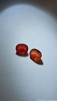 Pair of oval natural sapphires on white surface, showing deep orange with pink tones, faceted with inclusions.