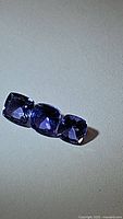 Photo showing three natural Tanzanite gemstones in cushion cut, blue-purple hue, arranged in a row on a plain surface.