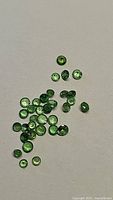 Scattered small round brilliant cut tsavorite garnet gemstones on a white background. Several dozen individual stones showing bright green color and varying sizes.