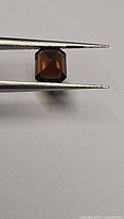 Close-up photo of the deep cognac orange-yellow octagon cut tourmaline gemstone held by tweezers showing facets and deep color.