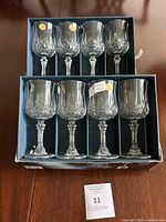 Full view of all 8 lead crystal wine glasses arranged in original compartmentalized box on wooden surface. The box shows some wear, glasses are clear with cut and faceted detailing.