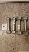 Four silver hex dumbbells arranged side by side on a wooden floor, showing weights and condition.