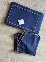 Flat lay of folded navy blue cloth placemats and napkins with gold border trim on a light wood floor.