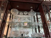 Crystal glassware displayed inside a cabinet on two glass shelves including bowls, vases and assorted dishes all with cut or etched patterns.