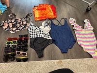 Photo showing four women's bathing suits arranged on the floor with ten pairs of sunglasses beside them.