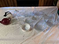 Wide view photo of assorted serving ware including glass bowls, crystal dishes, teapots, and glass cups laid out on white cloth.