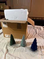 Three ceramic Christmas tree figurines in green, blue, and gray, displayed outside of the box on a table with the open cardboard box and white foam packaging visible behind them.