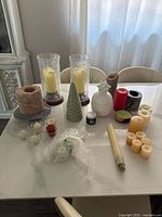 Full overview of all candle decor items on white table including glass hurricane holders, ceramic pineapple jar, pine tree decoration, assorted candles in various colors and sizes.