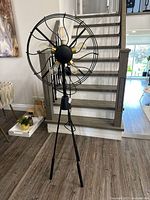 Front angled view of the vintage floor fan showing the brass-colored blades, cage, and tripod stand on wood flooring near stairs.