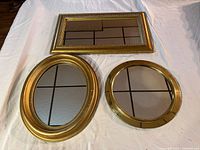 Three gold framed mirrors laid out flat on a white cloth, showing the rectangular, oval, and round shapes.