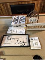 Photo showing assortment of home decor signs and wall art including windmill style sign, kitchen sign, 'love more worry less' framed art, house-shaped 'home' decor, welcome sign, and small block 'home' sign