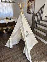 Kids white canvas teepee play tent with window and tied wooden poles indoors near stairs and dining area
