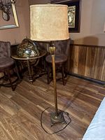 Photo of a vintage floor lamp with a light brown fabric drum shade, brass-tone metal stand and base, placed on a wooden floor near chairs.