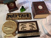 Front view of assorted man cave items on white surface showing decorative clock, green glass figurine, man cave sign, book-shaped box, miniature pistols display, bird plate, and chain with hooks.