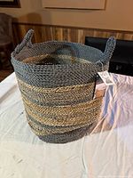 Front view of the new woven storage basket placed on a white cloth, showing its round shape, two handles, and alternating gray and natural color stripes.