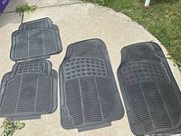 Four universal black rubber car mats laid outdoors on concrete and grass, showing full set including two larger front and two smaller rear mats with textured tread and anchor holes.