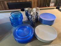 Large set of outdoor serving ware including blue translucent bowls, blue plastic plates, white textured plates, and boxed sets of blue-handled outdoor flatware, displayed on a pool table.