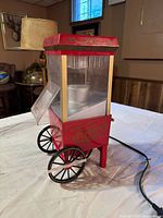 Red vintage style popcorn maker showing front and side clear panels with metal kettle inside and black spoked wheels.