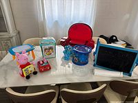 Full view of assortment of children's toys including Peppa Pig plush, Blue's Clues toy, red chair toy, drawing board, and chalkboard on a white table.