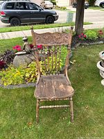 Photo shows a vintage wooden chair outdoors on grass. The chair has a carved floral scroll pattern on the backrest, spindles, and wear on the seat surface.