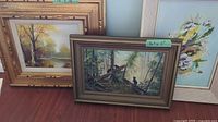 Framed paintings leaning against a wall on a wooden floor. One features bears in a forest, the other a landscape.