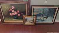 Three framed oil paintings arranged on floor: large pink rose floral, medium winter cabin scene, small snow river landscape
