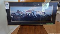 Framed panoramic numbered print artwork showing a mountain range with snow and peaks under a blue sky, placed on wooden floor.