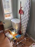 Photo showing ironing board, laundry basket, broom with dustpan, wooden shoe rack with hangers, cleaning cloths, and other accessories arranged in corner of room.