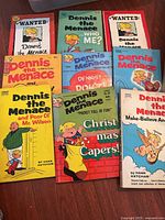 Close-up of multiple Dennis the Menace comic books with colorful vintage covers featuring the character and titles such as 'Wanted', 'Christmas Capers', and others.