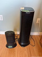 Photo of a Honeywell air purifier fan approximately 27 inches tall and a smaller Honeywell space heater fan approximately 13.5 inches tall, both plugged in and positioned on the floor against a wall.