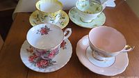 Four fine china teacups with matching saucers displayed on a wooden surface, showing floral patterns and gold trim.