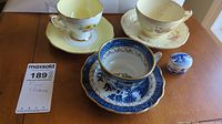 Photo showing three fine china tea cups with matching saucers and a small covered jar, arranged on a wooden table.