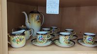 Full view of the china tea set including teapot, four teacups with saucers, creamer, and sugar bowl displayed on shelf.