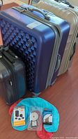 Three clam style suitcases in cream, blue and dark colors with luggage accessories on top of turquoise neck pillow.