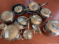 Overview of stainless steel pots and pans including lids, steamer, and wok on hardwood floor.